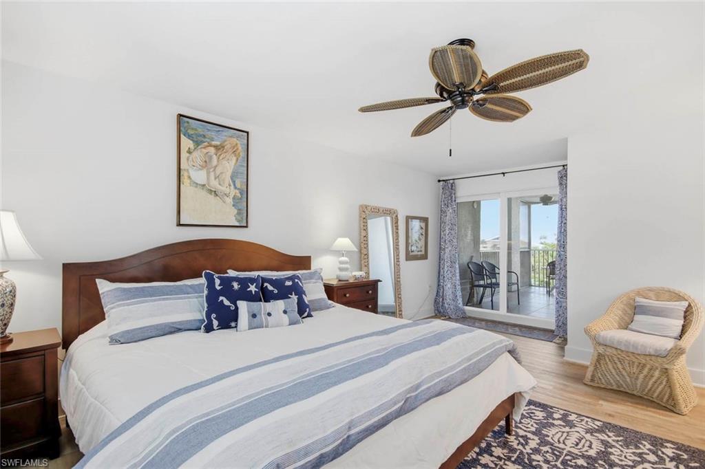 Primary bedroom featuring a wood-finish curved headboard, ceiling fan with brushed metal finish, sliding glass doors opening to a private balcony, wood-finish flooring, and an en-suite bathroom with framed mirror and vanity