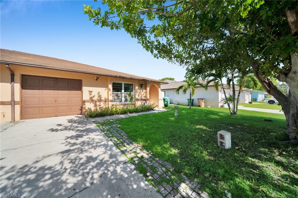 Charming single-story residence featuring a wood-finish garage door, manicured front lawn with mature shade trees, and concrete driveway with decorative brick edging
