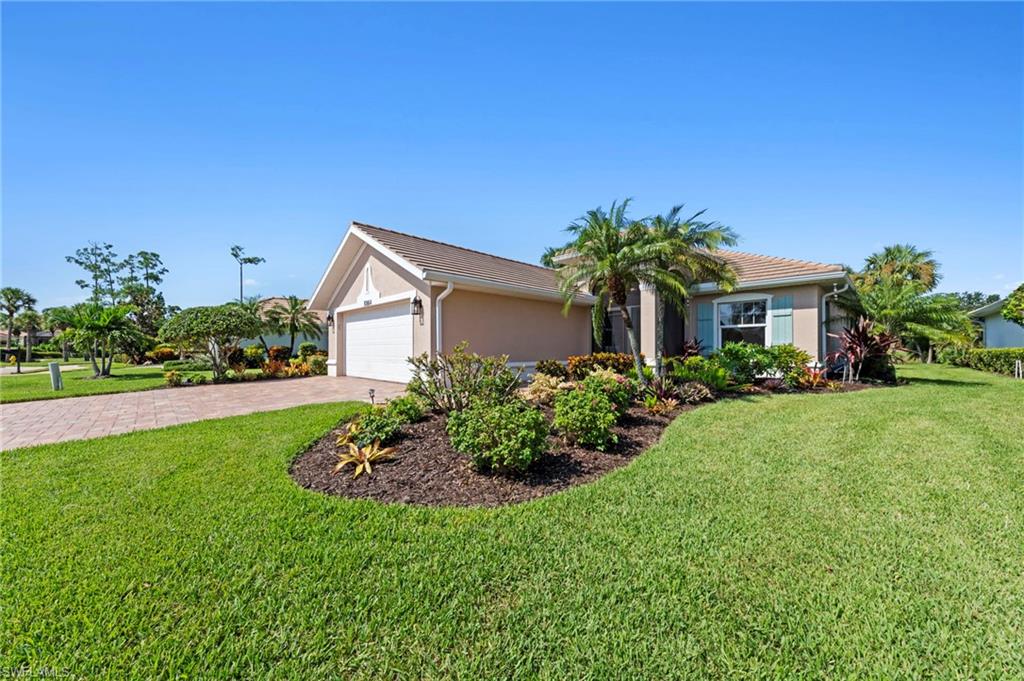 Charming single-story residence featuring a pristine manicured lawn, mature landscaping with tropical palms and flowering shrubs, brick paver driveway, and a two-car garage with white panel doors
