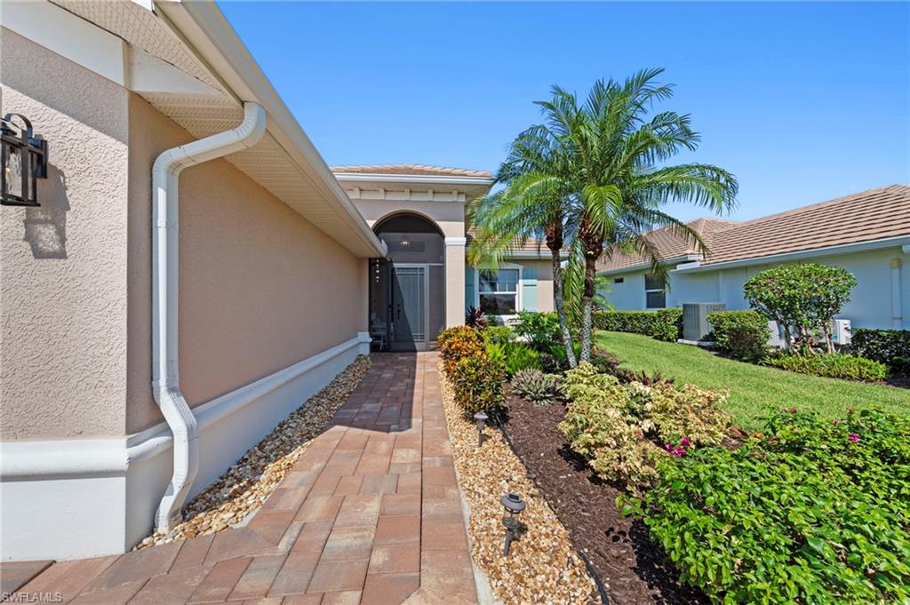 Welcoming entry courtyard featuring a brick paver walkway, mature palm tree, manicured landscaping with low hedges and tropical plantings, and stucco exterior with white trim detailing