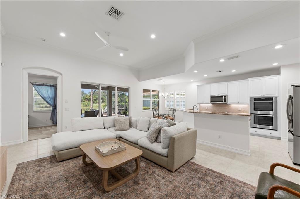 Open-concept living and kitchen featuring vaulted ceilings with recessed lighting, large multi-pane windows, light wood-finish flooring, neutral cabinetry with integrated appliances including dual wall ovens, stainless steel refrigerator, and a wood-top island with seating