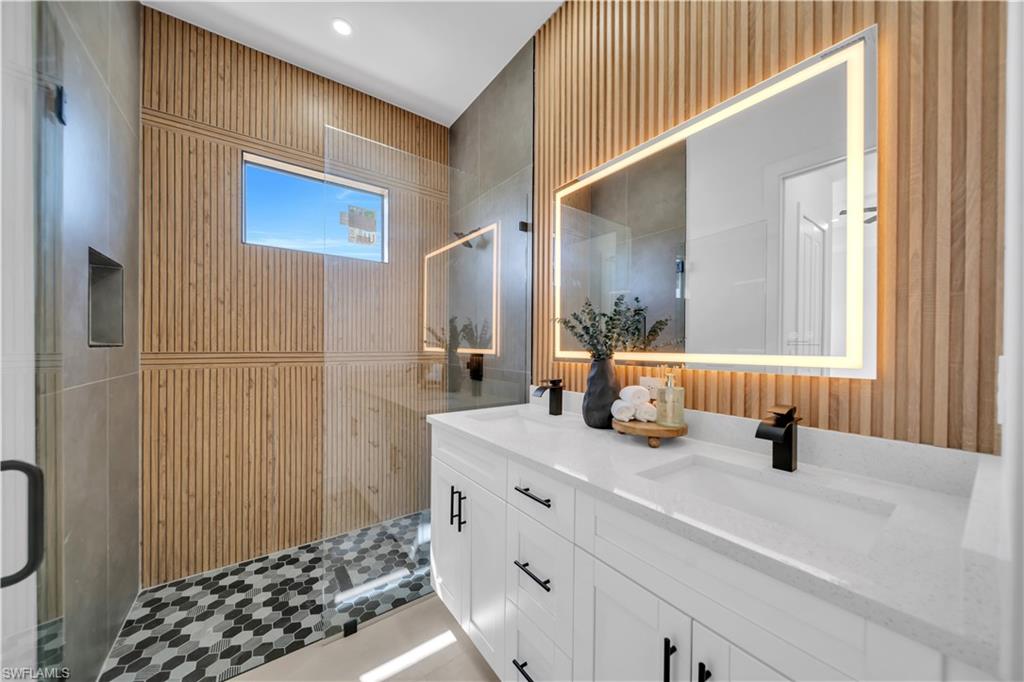 Double vanity with integrated sinks and black matte faucets, wood-finish slatted accent wall, rectangular backlit mirror, glass-enclosed shower, and hexagonal mosaic shower flooring