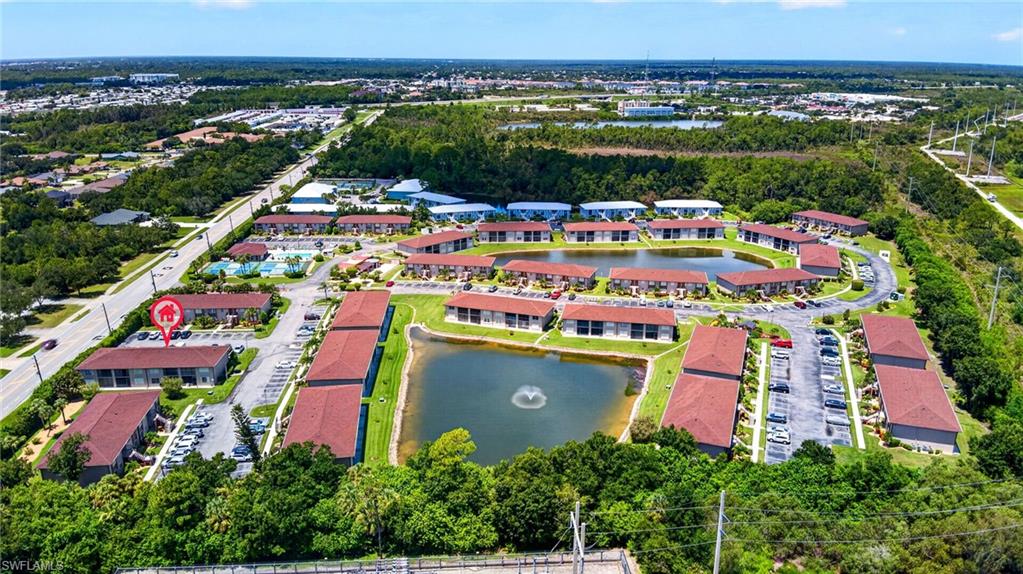Community setting with multiple buildings featuring tiled roofs, surrounding a central pond with a fountain, and ample surface parking