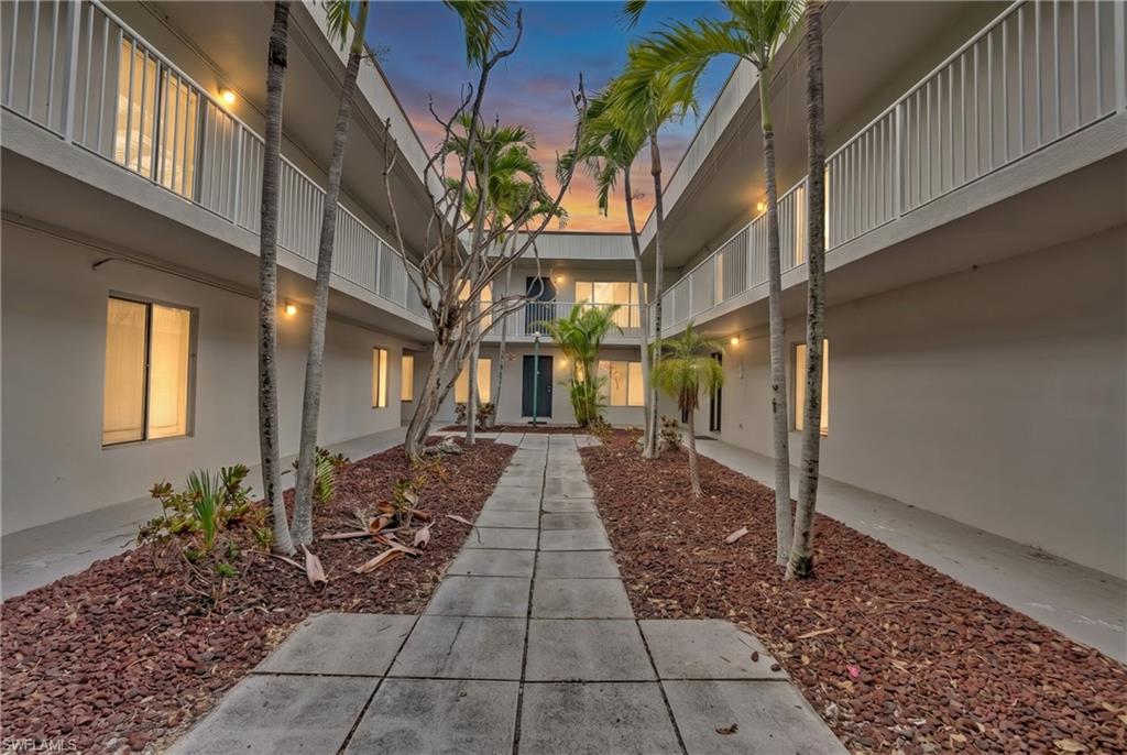 Two-story building exterior with multiple balconies featuring white railings, surrounded by mature palm trees, and a central walkway with red rock landscaping