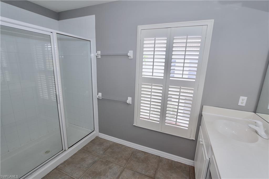 Glass-enclosed shower with white frame, white vanity with integrated sink, white plantation shutters, and large format tile flooring