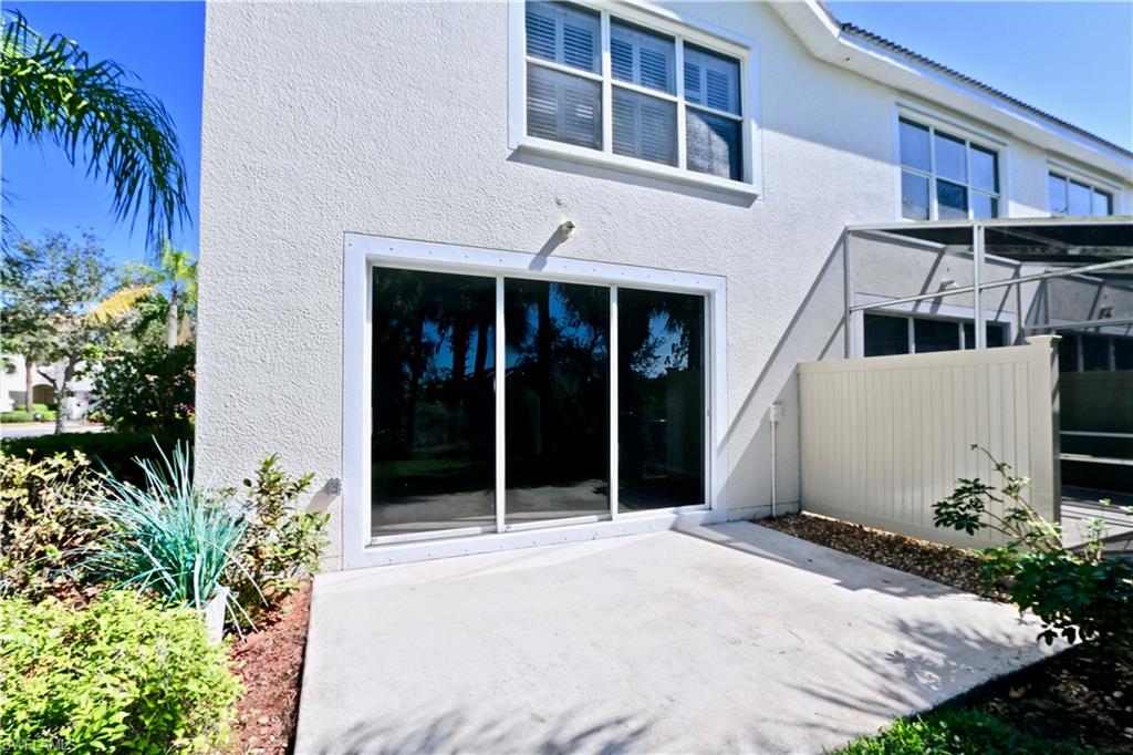 Exterior wall with stucco finish, ground-level concrete patio, sliding glass door, white window frames with horizontal blinds, and mature landscaping
