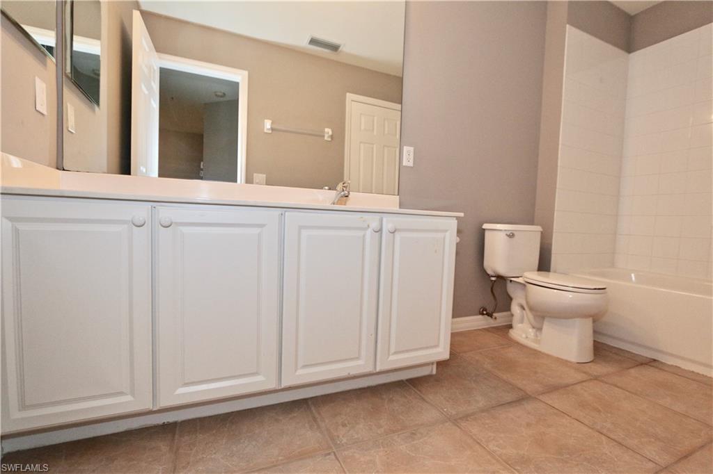 Bathroom featuring a white vanity with panel cabinetry, a built-in tub with white tile surround, a toilet, and neutral-toned tile flooring