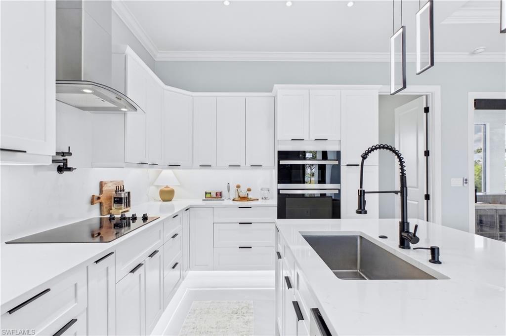 White shaker cabinetry with black hardware, integrated built-in oven, stainless steel range hood, sleek cooktop, and an island with an undermount sink and black faucet