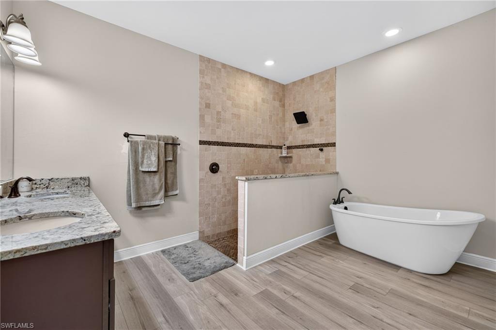 Freestanding soaking tub, walk-in shower with tiled walls, dual vanity with undermount sink, recessed lighting, and wood-finish flooring