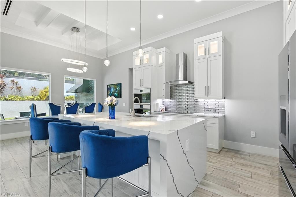 Kitchen with wall chimney exhaust hood, crown molding, tasteful backsplash, a center island with sink, and light stone countertops
