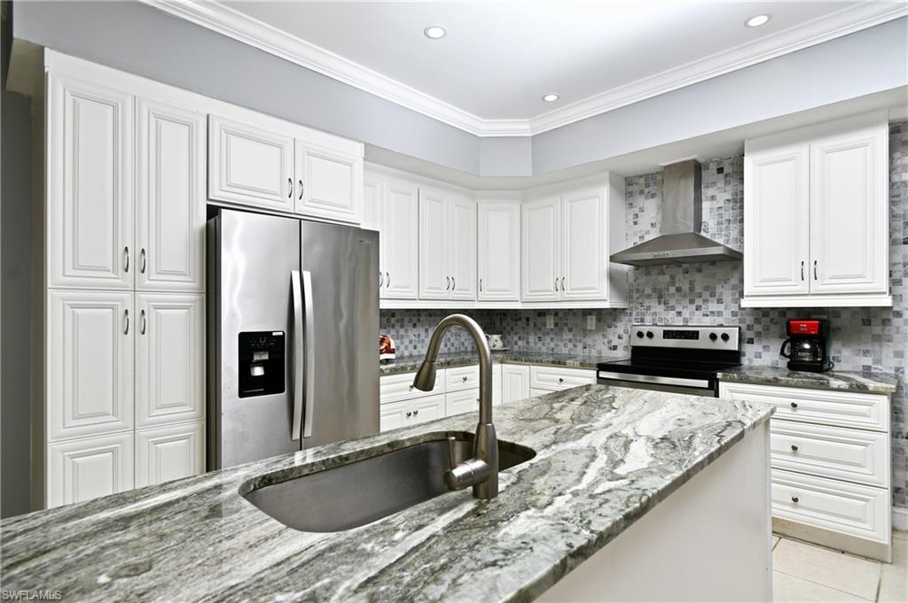 Kitchen with white paneled cabinetry, stainless steel appliances, mosaic tile backsplash, and a large island featuring a built-in sink and stone countertop