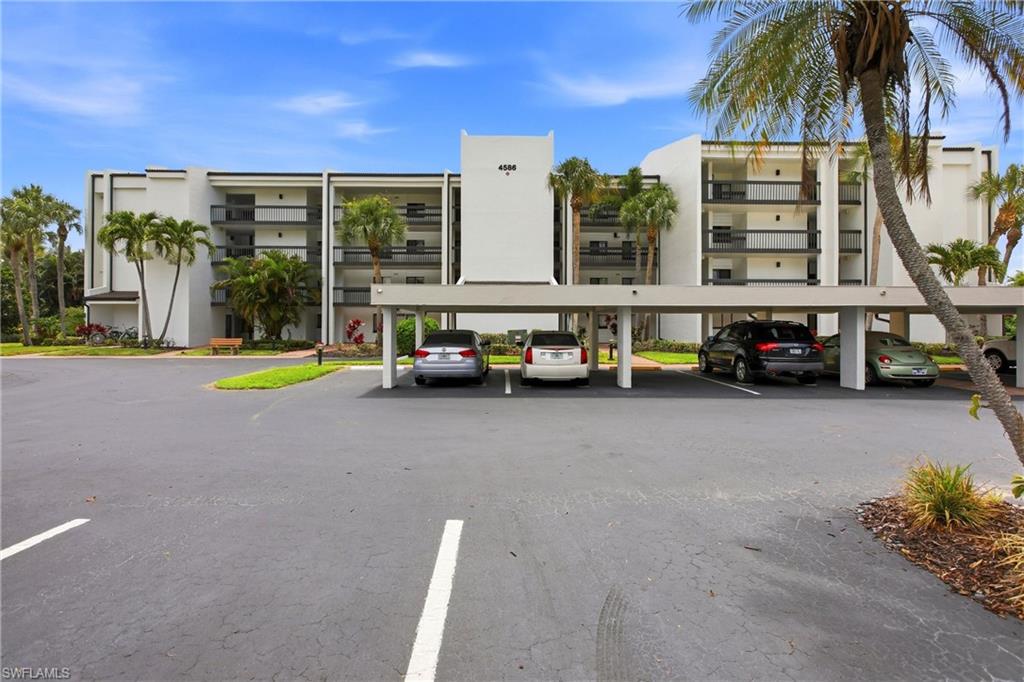 Contemporary multi-story building featuring covered parking, multiple balconies with dark railings, a light-colored exterior, and surrounding tropical landscaping