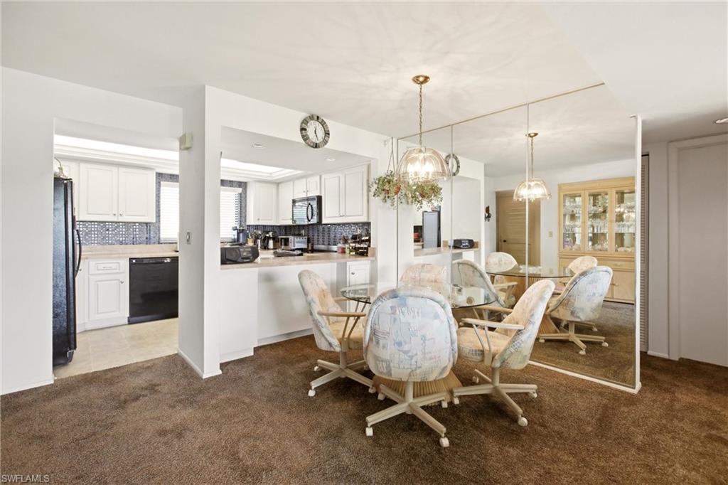 Dining area with full-wall mirror paneling, a contemporary chandelier, and brown carpeting, opening to a kitchen featuring white cabinetry, black appliances, and a tiled backsplash