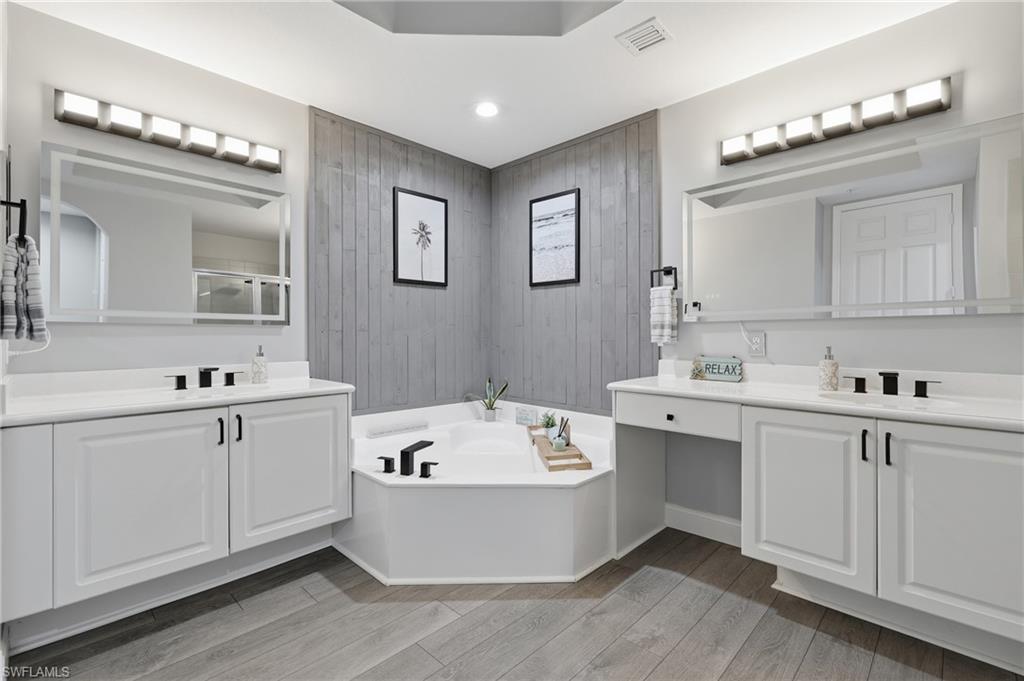 Dual vanities with white cabinetry, corner soaking tub, plank-style flooring, and wall-mounted light fixtures