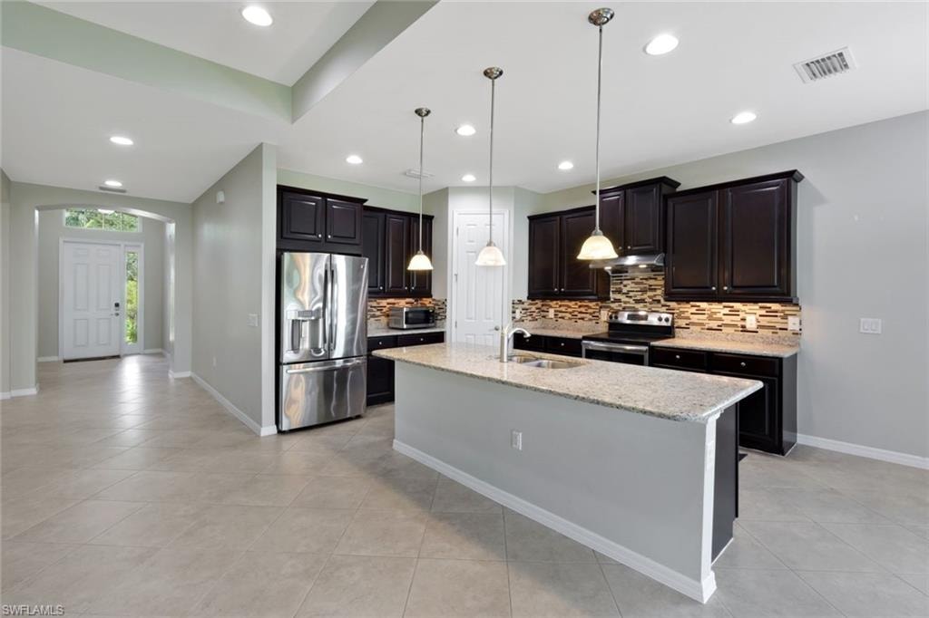 Kitchen featuring dark wood cabinetry, stainless steel appliances, a multi-tone tile backsplash, and an island with a light-toned countertop