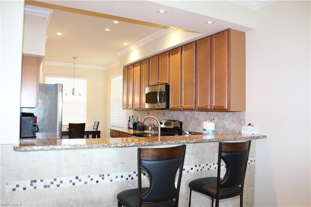 Kitchen featuring ornamental molding, tasteful backsplash, light stone counters, decorative light fixtures, and brown cabinetry