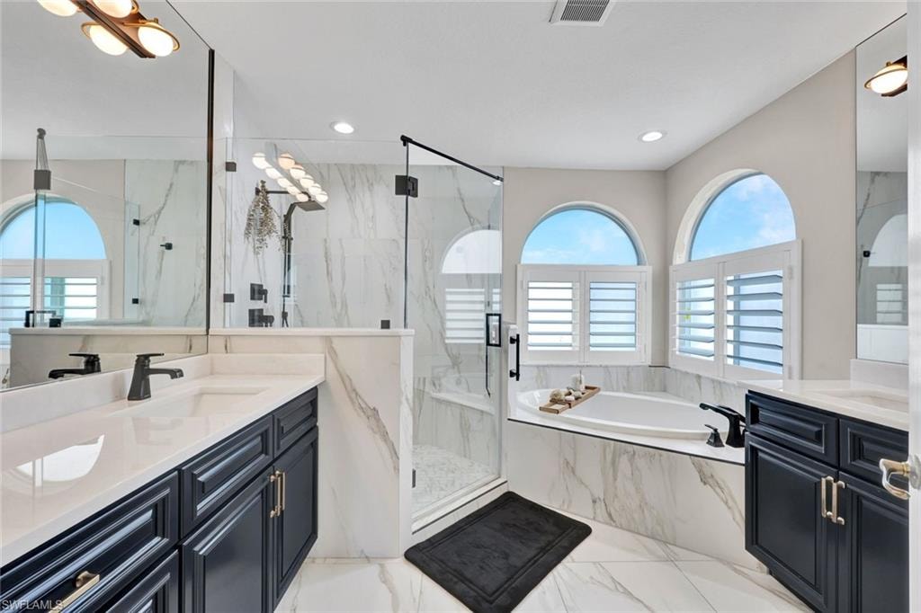 Dual vanities with undermount sinks, a built-in soaking tub, an enclosed glass shower, and arched windows with fogged glass and plantation shutters