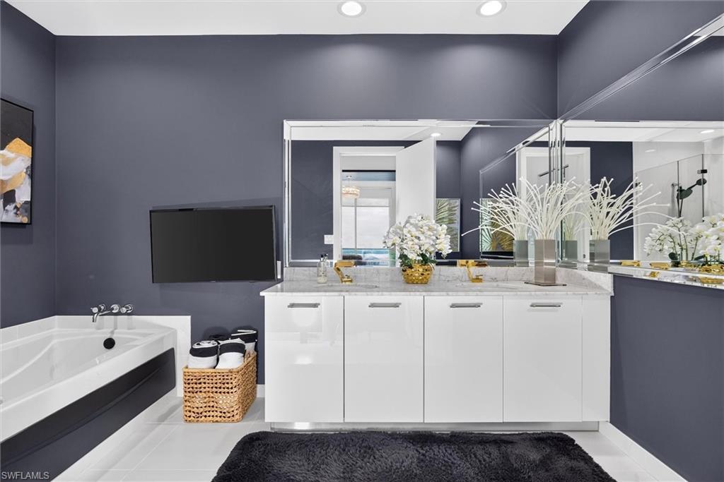 Dual vanity with a stone countertop, wall-mounted television, built-in bathtub, recessed lighting, and wall-mounted mirrors