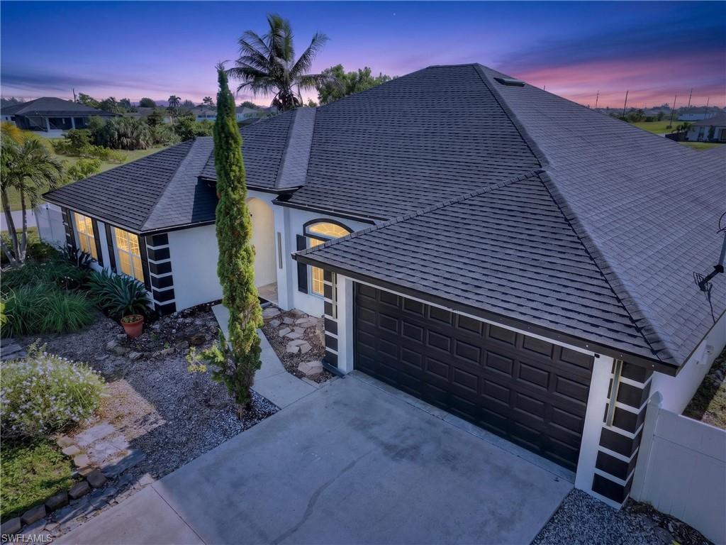 Pitched shingle roof, white stucco exterior with dark trim accents, attached garage with panel door, concrete driveway, and established front landscaping