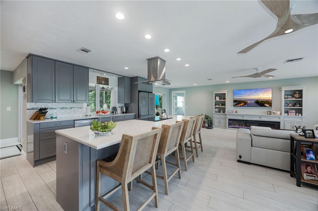 Spacious open-concept living area featuring a kitchen island with seating, gray shaker cabinetry, stainless steel appliances, and a built-in entertainment center with an electric fireplace