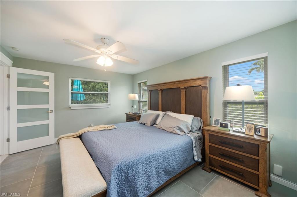Wood-finish bed frame, wood-finish nightstands, ceiling fan with integrated lighting, multiple windows with blinds, and large-format tile flooring