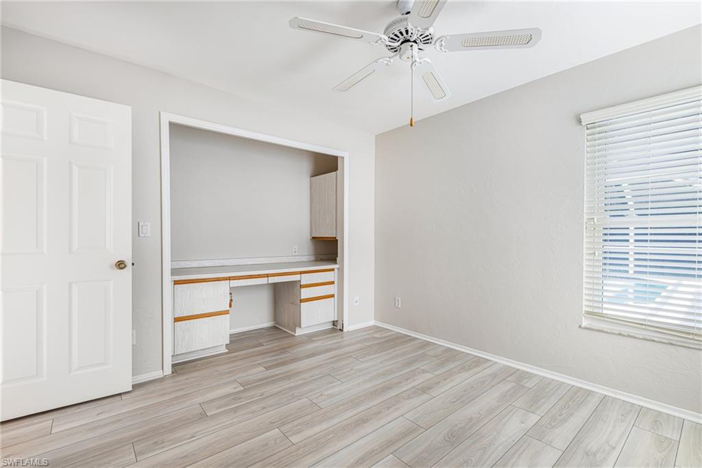 Light wood-finish flooring, built-in desk with cabinetry, window with blinds, and a ceiling fan