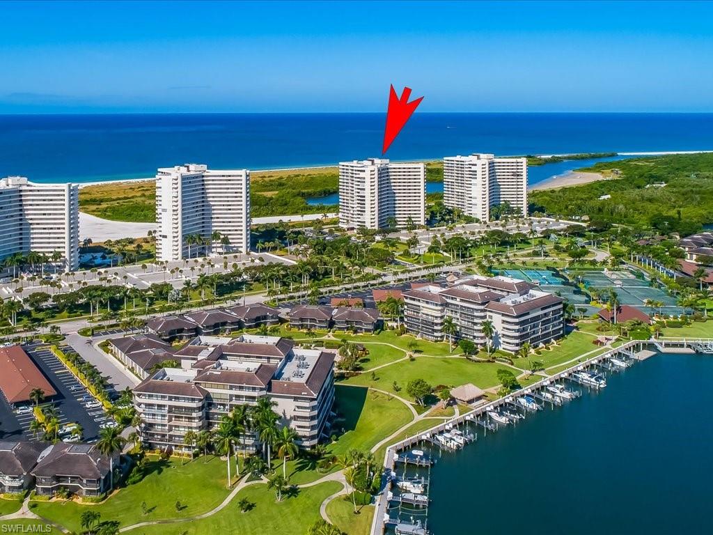 Coastal high-rise buildings with balconies, extensive green spaces, a marina with numerous boat slips, multiple tennis courts, and direct proximity to a sandy coastline and water