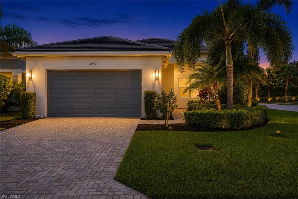 Paver driveway, exterior wall sconces, integrated garage, manicured shrubbery, and mature palm trees