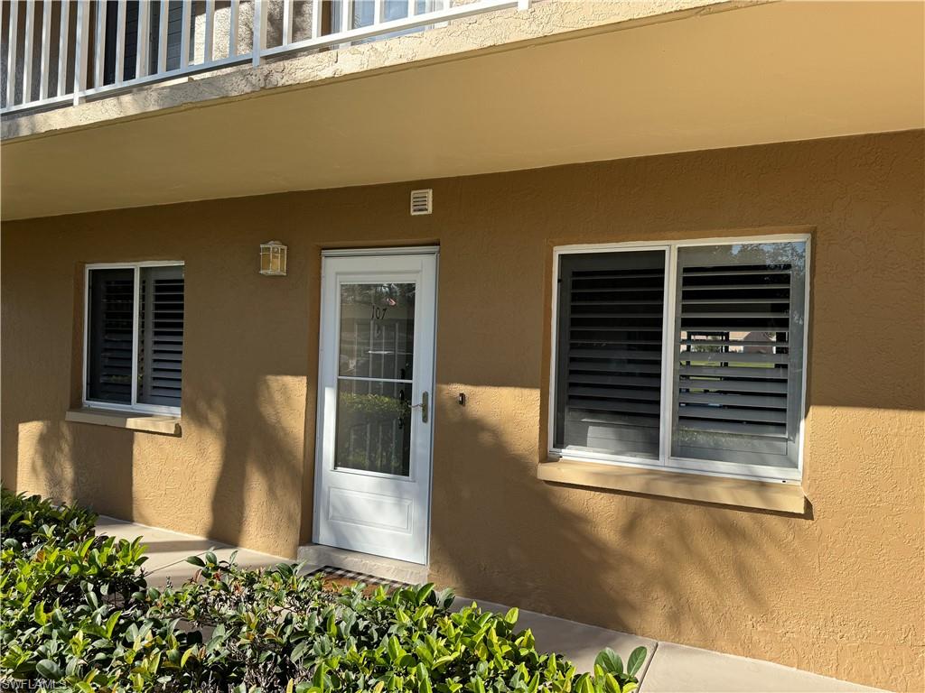 Stucco exterior with a white entry door, exterior sconce lighting, and white-framed windows with louvered shutters