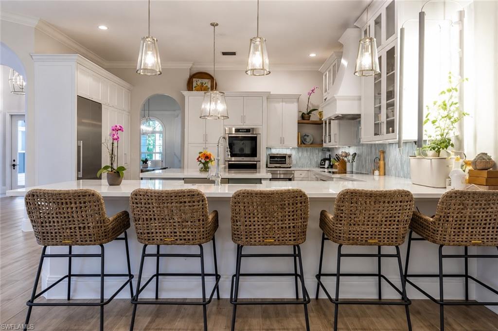 Expansive kitchen island with seating, white cabinetry, integrated stainless steel refrigerator, built-in oven, and pendant lighting