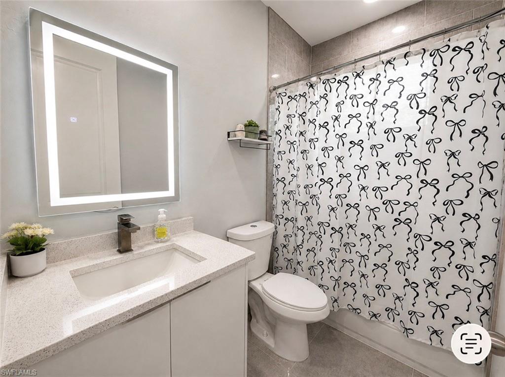 Illuminated vanity mirror, single-basin vanity with a light-toned countertop, dark-toned faucet, white vanity cabinetry, and tiled shower surround