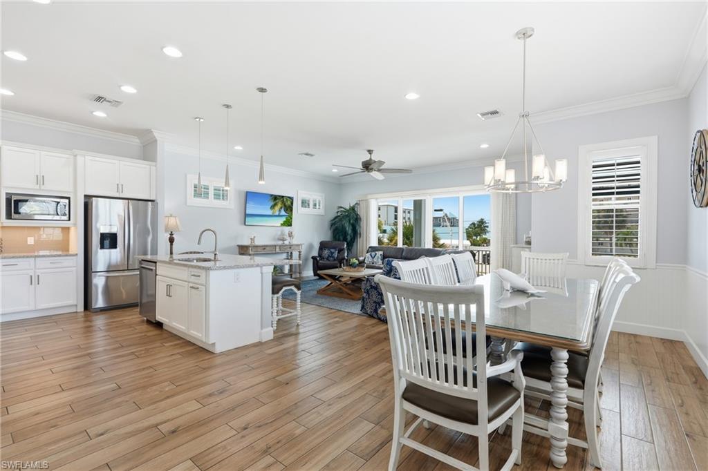 Open-concept living area with wood-finish flooring, a kitchen island with seating, stainless steel appliances, white cabinetry, and a dining area with a chandelier - Virtually Edited Image