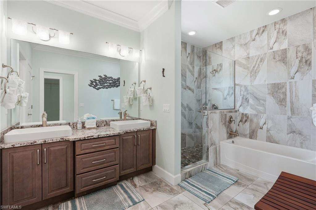 Dual vessel sinks set atop a granite-finish countertop, a glass-enclosed shower with pebble-finish flooring, a soaking tub, and floor-to-ceiling tile in the shower and tub surround - Virtually Edited Image