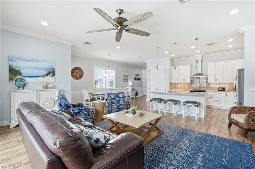 Open-concept living area with wood-finish flooring, recessed lighting, a ceiling fan, and white kitchen cabinetry with a central island - Virtually Edited Image