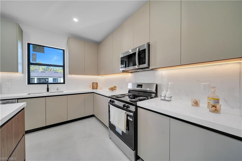 Modern kitchen with white countertops, stainless steel appliances, light-toned cabinetry, under-cabinet lighting, and a black-framed window