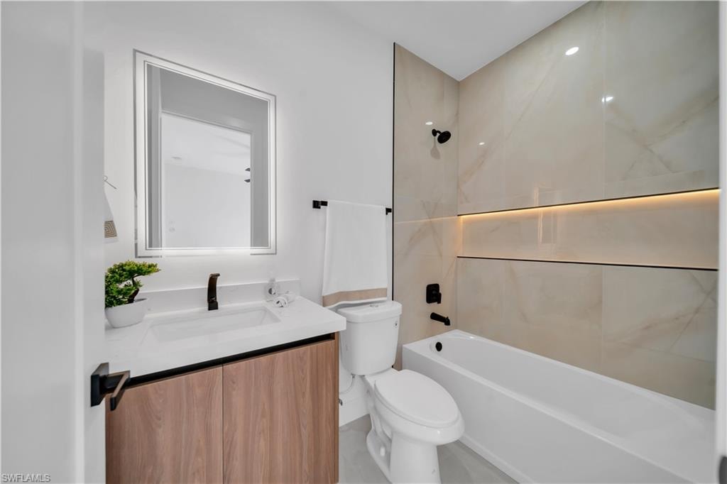 Bathroom featuring a built-in bathtub, light wood-finish vanity, illuminated mirrored medicine cabinet, matte black fixtures, and tiled shower surround with accent lighting