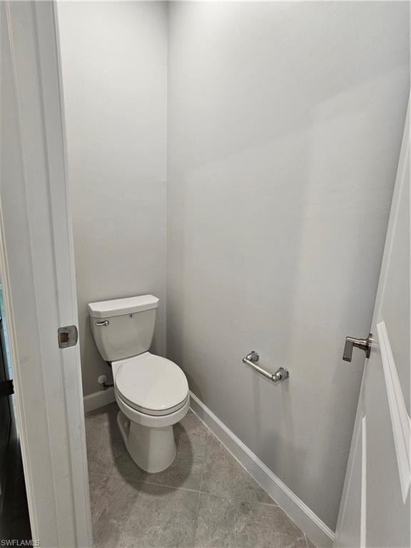 White toilet with a round bowl, gray floor tiling, chrome toilet paper holder, white baseboards, and a white door with a satin nickel lever handle