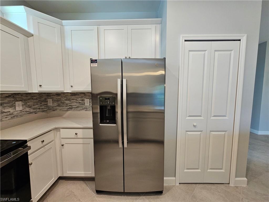 Stainless steel side-by-side refrigerator, white upper and lower cabinetry, mosaic tile backsplash, solid surface countertops, and tile flooring