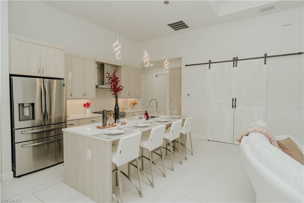 Kitchen with decorative light fixtures, visible vents, light countertops, stainless steel refrigerator with ice dispenser, and a center island with sink