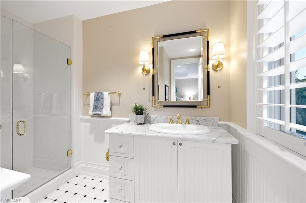 Glass-enclosed shower, white beadboard wainscoting, white vanity with stone countertop, polished brass fixtures, and a black and white tiled floor