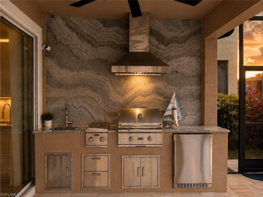 Outdoor kitchen featuring a stone accent wall, built-in stainless steel grill, integrated range hood, under-counter refrigeration, and a ceiling fan