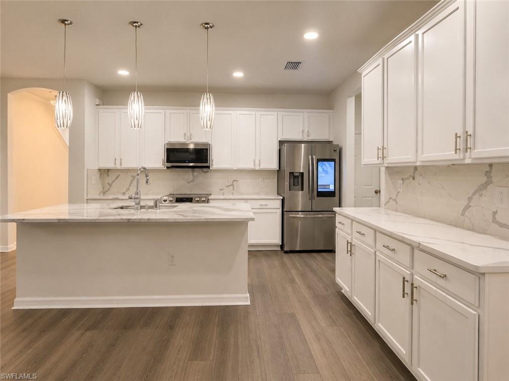 White cabinetry with gold-tone hardware, stainless steel appliances, wood-finish flooring, island with integrated sink, and recessed lighting