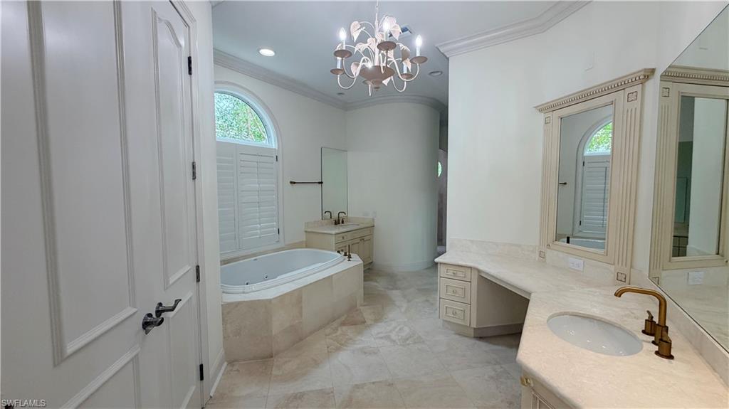 Spacious bathroom featuring a built-in jetted tub, arched window with plantation shutters, dual vanities with integrated sinks, and a decorative chandelier