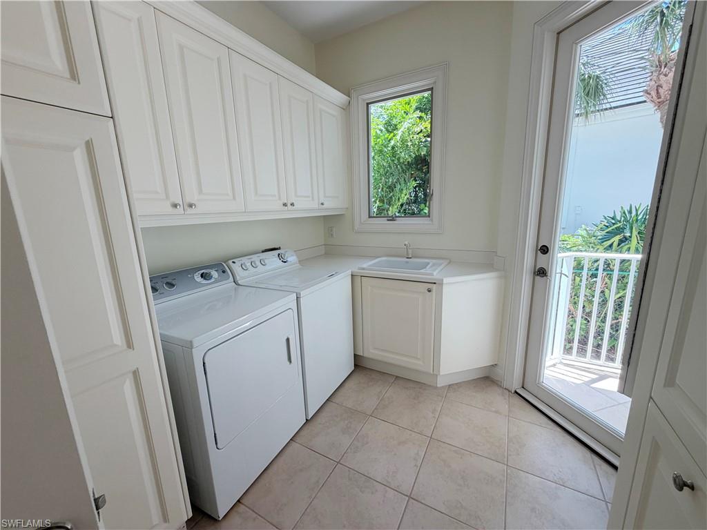 Dedicated laundry room featuring white cabinetry with ample storage, stacked washer and dryer appliances, tile flooring, utility sink with cabinet base, and abundant natural light from dual glass doors and window overlooking outdoor space