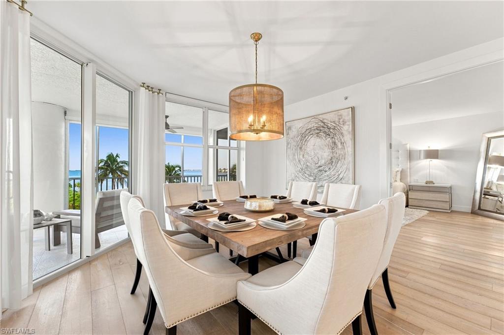 Open-concept dining area with wood-finish flooring, a contemporary chandelier, and a wide balcony with water and palm tree scenery