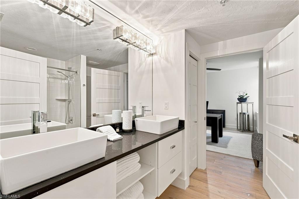Double vessel sinks, dark countertop, white shaker-style cabinetry, chrome fixtures, and wood-finish flooring