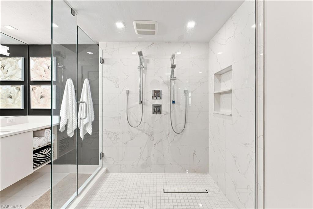 Glass-enclosed shower with dual shower heads, white marble-look wall tiles, white square floor tiles, and a recessed shower niche