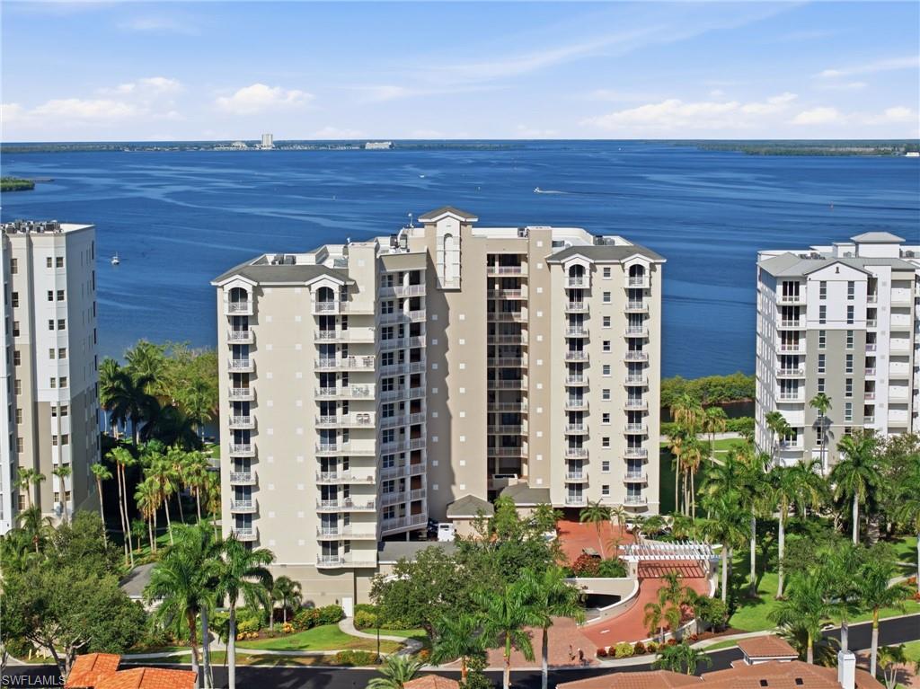 High-rise condominium with multiple balconies, a structured portico entrance, lush landscaping, and a water backdrop
