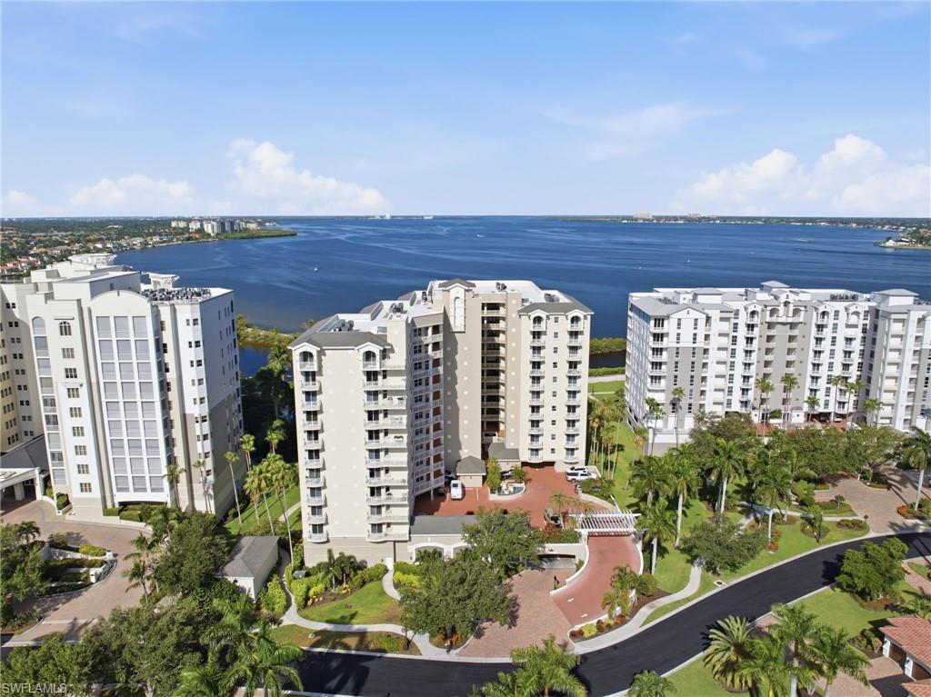 High-rise condominium structures with multiple balconies, surrounded by mature landscaping, and featuring a brick paved entry drive, all situated along a substantial body of water