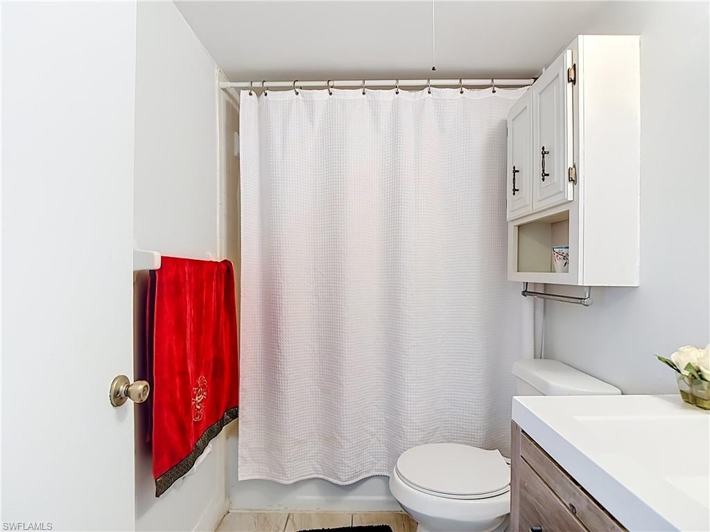 Bathroom featuring a white shower curtain, white wall cabinet with dark hardware, toilet, and a wood-finish vanity with a white countertop and integrated sink